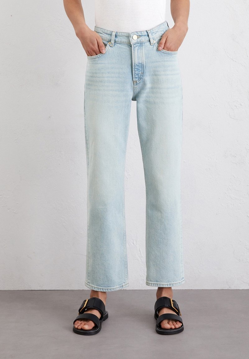 Persona con jeans rectos de color azul claro y sandalias negras con hebillas doradas, de pie contra una pared blanca texturizada.