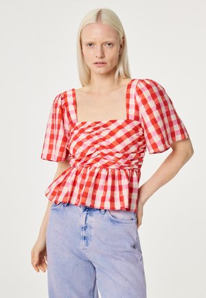 Vrouw met steil blond haar, gekleed in een rood- en roze geruit blouse met pofmouwen en lichtblauwe jeans met hoge taille, poseert met hand op de heup.