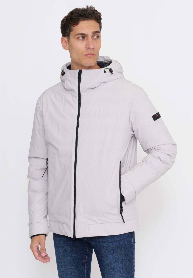 Uomo che indossa una giacca con cappuccio di colore grigio chiaro con cerniera e tasche nere, in piedi e che guarda di lato su uno sfondo semplice.