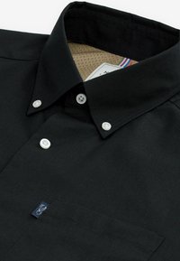 Schwarzes Hemd mit Knopfleiste, Kragen und Tasche; es verfügt über weiße Knöpfe, strukturierte Stoffqualität und ein gepunktetes Innenfutter mit einem Etikett.