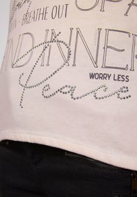 T-shirt in cotone rosa chiaro con decorazioni in strass argentati e design testuali. L'orlo è leggermente curvo con una cucitura visibile.