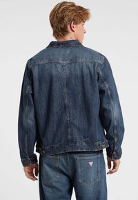 Giubbotto di jeans blu scuro con un taglio classico. Presenta un colletto, chiusura con bottoni e tasche sul petto. La texture mostra una finitura vissuta.