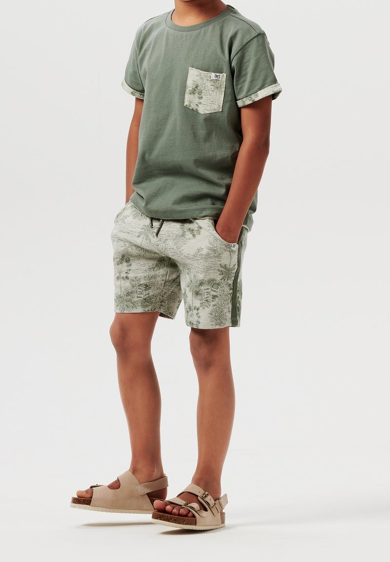 Enfant portant un t-shirt vert à manches courtes avec une poche à motif, un short imprimé assorti et des sandales beiges à boucle, debout les mains dans les poches.