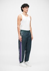 Dunkelgrüne Sporthose mit weißen und lila Seitenstreifen, elastischem Bund und Reißverschlusstaschen. Kombiniert mit einem weißen Tanktop und weißen Sneakern.