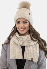 Gorro de punto crema con un pompón esponjoso y bufanda a juego que presenta un patrón texturizado y bordes cosidos. Acento de piel marrón en la bufanda.