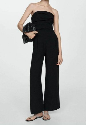 Frau trägt trägerlosen schwarzen Jumpsuit mit weitem Bein, hält eine schwarze geflochtene Clutch und trägt flache Sandalen mit transparenten Riemen.