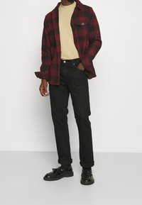 Rood-zwart geruit flanellen overhemd, beige T-shirt met ronde hals, zwarte spijkerbroek, zwarte schoenen, tentoongesteld staand tegen een effen achtergrond.