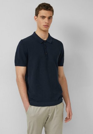 Młody mężczyzna stoi z rękami w kieszeniach, ubrany w granatową, teksturowaną koszulkę polo z krótkim rękawem oraz beżowe spodnie, na jasnym tle.