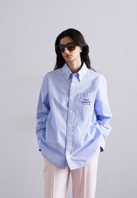 Heledad helesinise triibulise nööbiga särgi, millel on rinnatasku tekstiga "THE SHIRT". Kandmine on lõdva stiiliga, koos heledate roosa värvi pükstega.