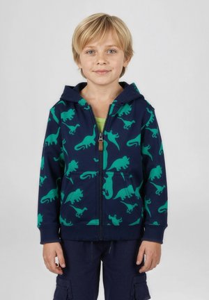 Bambino biondo con felpa con cappuccio blu navy con stampe di dinosauri verdi e pantaloni cargo blu navy, in piedi davanti a uno sfondo chiaro uniforme.