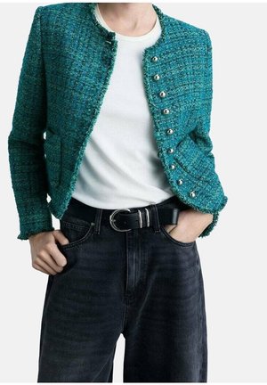 Kort, tekstureret jakke i blågrøn med sølvknapper, båret over en hvid top og sort bælte med sølvspænde, kombineret med mørke, højtaljede jeans.