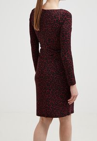Femme aux longs cheveux portant une robe noire ajustée à manches longues, arrivant au niveau des genoux, avec un motif floral abstrait rouge, vue de dos.