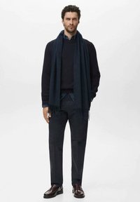 Suéter de canalé azul marino, capas con una bufanda oscura, combinado con pantalones oscuros y mocasines marrones. El modelo se encuentra de pie contra un fondo sencillo.