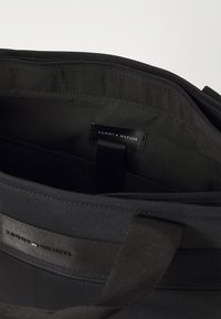 Sac fourre-tout en tissu noir avec fermeture éclair ; doublure intérieure en gris foncé ; possède une étiquette Tommy Hilfiger et des poignées robustes.