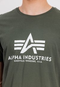 Olivgrünes Baumwoll-T-Shirt mit einem weißen Logo, das stilisierte Buchstaben und parallele Linien zeigt, Rundhalsausschnitt und glatter Textur.