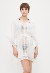 Valge kaftan, mis on valmistatud õhukesest kangast, millel on peen pitsdetail kaelaosas ja hemper, ning see omab lõdva ja voogava siluetti.