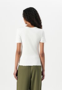 Top bianco a maniche corte con costine, orlato in modo scallop, abbinato a pantaloni verdi a vestibilità ampia, che evidenziano un design e una texture casual.