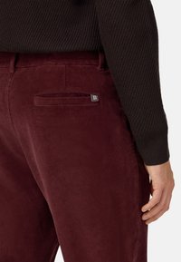 Bordeaux cordflers med en bakficka och en liten märkesetikett. Tyget har en ribbad textur, kombinerad med en mörk stickad tröja.