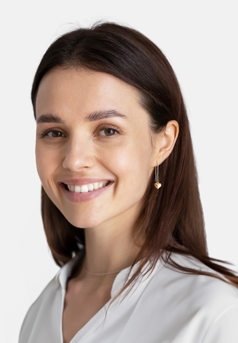 Goldene herzförmige Ohrringe mit einer zarten Kette, getragen von einem Model. Das Model hat einen glatten Teint und lange dunkle Haare, bekleidet mit einer weißen Bluse.