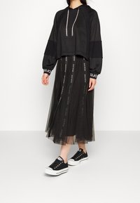 Czarna krótka bluza z kapturem o fakturowanym wykończeniu i przodem ze sznurkiem, zestawiona z czarną spódnicą z tiulu z akcentami z taśmy z logo oraz sneakersami na platformie.