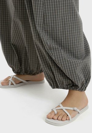 Voeten met witte platte sandalen met bandjes, gecombineerd met losse, donkere geruite broeken samengebonden bij de enkels op een witte achtergrond.