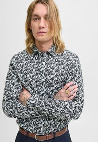 Juodas gėlių marškinėliai su baltais žiedais, ilgomis rankovėmis ir pritaikyta forma. Įsikišti į tamsias kelnes su rudomis diržais.