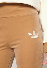 Hnědé sametové tepláky s bílým logem Adidas na levém stehně, s hladkou texturou a čistým designem.