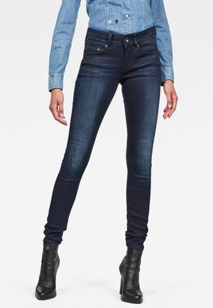 Jeans ajustados en denim azul oscuro con un ligero brillo. El diseño cuenta con un estilo clásico de cinco bolsillos y una cintura de altura media.