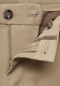 Tissu beige avec une braguette zippée, un bouton en métal et des détails de couture. L'intérieur présente une doublure marron contrastante.