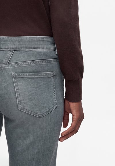 Gros plan d'une personne portant un jean gris délavé et un haut à manches longues marron foncé, montrant la poche arrière et la main droite détendue sur le côté.