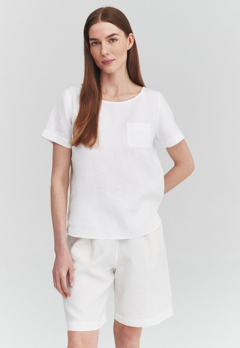 Femme aux longs cheveux bruns portant un haut blanc à manches courtes avec poche poitrine et un short blanc jusqu'aux genoux, debout devant un fond uni.