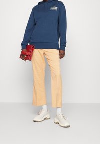 Felpa blu navy con logo, pantaloni cropped a quadri pesco, sneaker bianche con suole beige e una borsa a clutch rossa con texture.