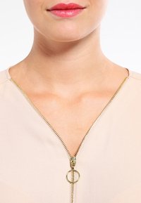 Blusa beige con scollatura a V e dettaglio della cerniera dorata. La cerniera presenta un tirante circolare e si estende dalla scollatura verso il basso.