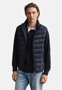 Marinblå dunväst med quiltad design, dragkedjefickor och hög krage, bärs över en mörk pikétröja. Blå jeans kompletterar looken.