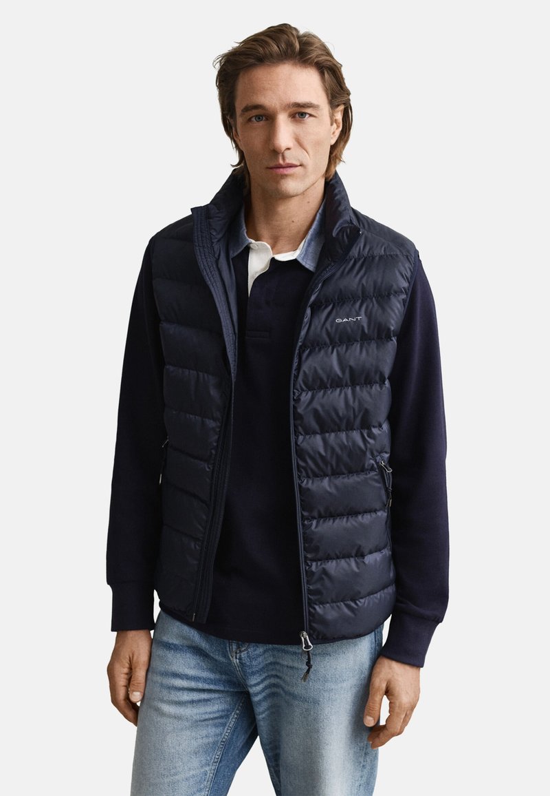 Marinblå dunväst med quiltad design, dragkedjefickor och hög krage, bärs över en mörk pikétröja. Blå jeans kompletterar looken.