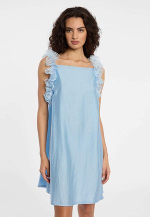 Femme aux cheveux bouclés portant une robe bleu clair sans manches avec des bretelles à volants, debout devant un fond blanc uni.