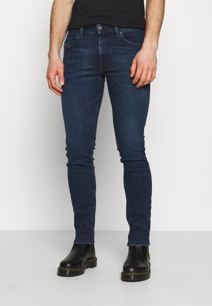 Homme portant un jean slim bleu foncé et des bottes en cuir noires, debout devant un fond blanc.