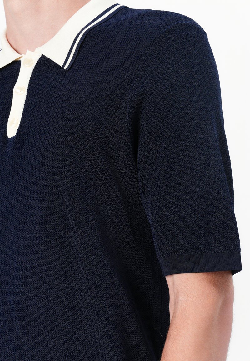 Navyblaues Polohemd mit kurzen Ärmeln aus strukturiertem Stoff, mit einem cremefarbenen Kragen mit navyblauen Streifen und einer Knopfleiste mit drei Knöpfen.