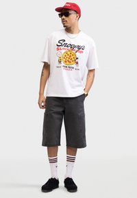 Hvid grafisk T-shirt med et pizzadesign og tegneseriefigurer. Mørkegrå cargo shorts, sorte sko og en rød cap med solbriller.