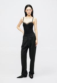 Schwarzer Body mit Spitzen-Details und dünnen Trägern, kombiniert mit hoch taillierter schwarzer Hose. Model posiert selbstbewusst vor neutralem Hintergrund.