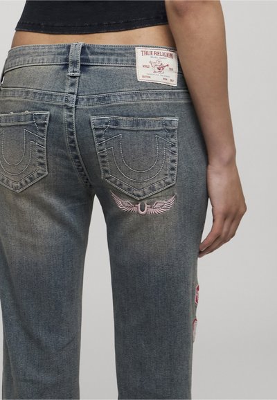Vue de dos d'une personne portant un jean True Religion bleu délavé avec un patch logo et une broderie sur la poche arrière droite.