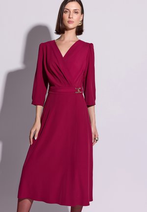 Fuksjowa sukienka kopertowa z rękawami do łokcia, zdobiona talią z pozłacaną klamrą paska i zwiewną spódnicą, wykonana z gładkiej tkaniny.