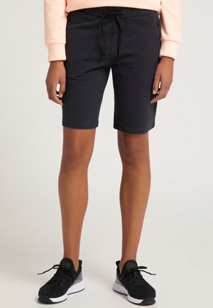 Personne portant un short noir au genou avec un cordon, un haut à manches longues couleur pêche et des chaussures de sport noires, debout sur un fond neutre.
