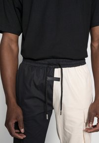 Puoli mustat ja puoli kermaiset collegehousut, joissa on joustava vyötärönauha, kiristysnauha, sivutasku ja "SIXTH JUNE" -logolla varustettu etiketti.