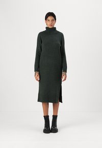 Abito in maglia di colore verde scuro con colletto alto, maniche lunghe, spacchi laterali e polsini a costine. Indossato con stivali neri con lacci.