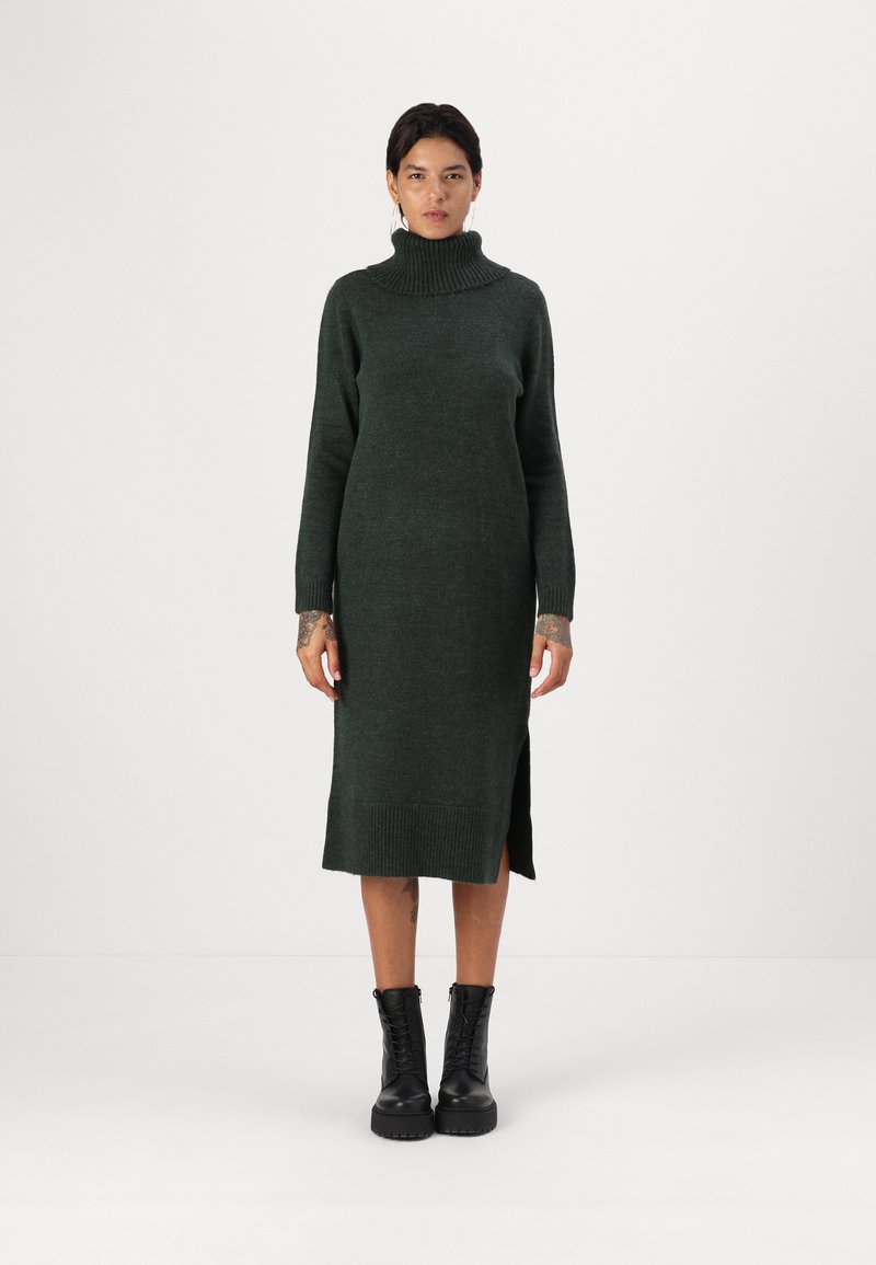 Abito in maglia di colore verde scuro con colletto alto, maniche lunghe, spacchi laterali e polsini a costine. Indossato con stivali neri con lacci.