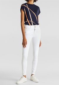 Marinblå topp med kort ärm och orangea och vita abstrakta linjer, kombinerad med vita slim-fit jeans och vita sneakers. Slät, lättviktig tyg.