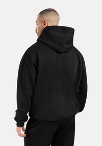 Sudadera negra de gran tamaño con capucha ajustable, puños acanalados y una textura suave, combinada con pantalones negros. Destacada por su corte holgado.