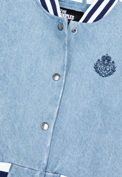 Jasnoniebieska kurtka dżinsowa z zatrzaskami, granatowym haftowanym herbem na piersi oraz ściągaczami przy kołnierzu i mankietach w granatowo-białe paski.