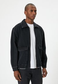 Hombre de pie vestido con una chaqueta negra con cremallera y puños elásticos sobre una camiseta blanca y pantalones negros, frente a un fondo plano.